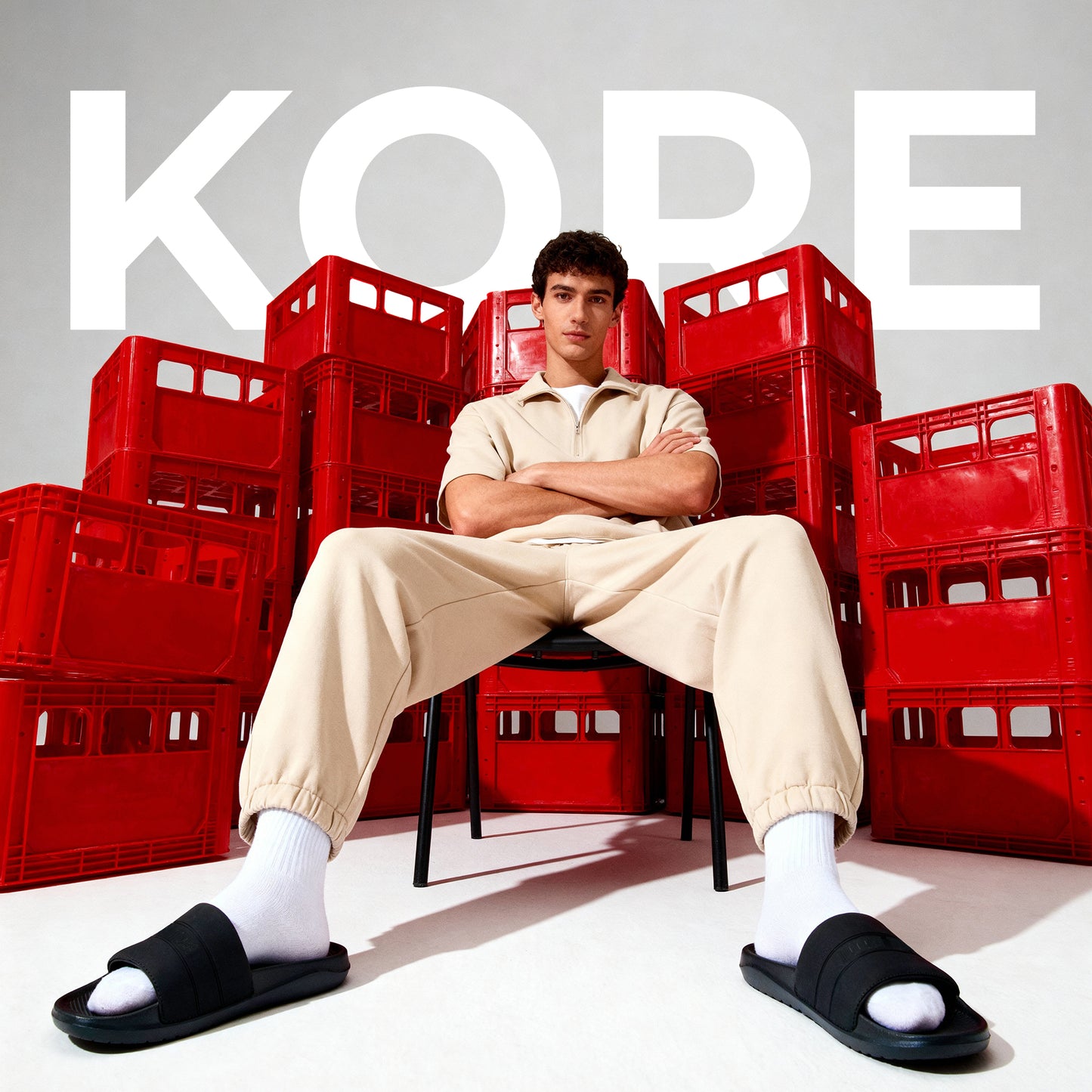 Male model wearing black slide sandals with white socks and beige casual outfit sitting against stacked red crates