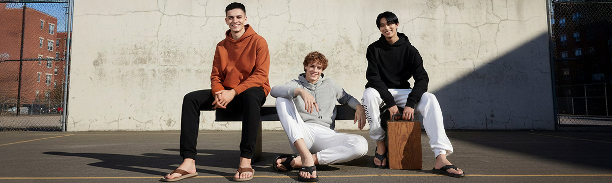 Three young men in casual hoodies and white joggers relaxing on urban basketball court on sunny day