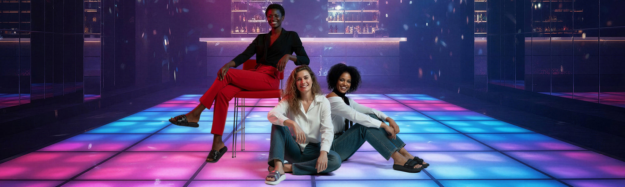 Three stylish women posing on a colorful illuminated dance floor in a modern bar lounge