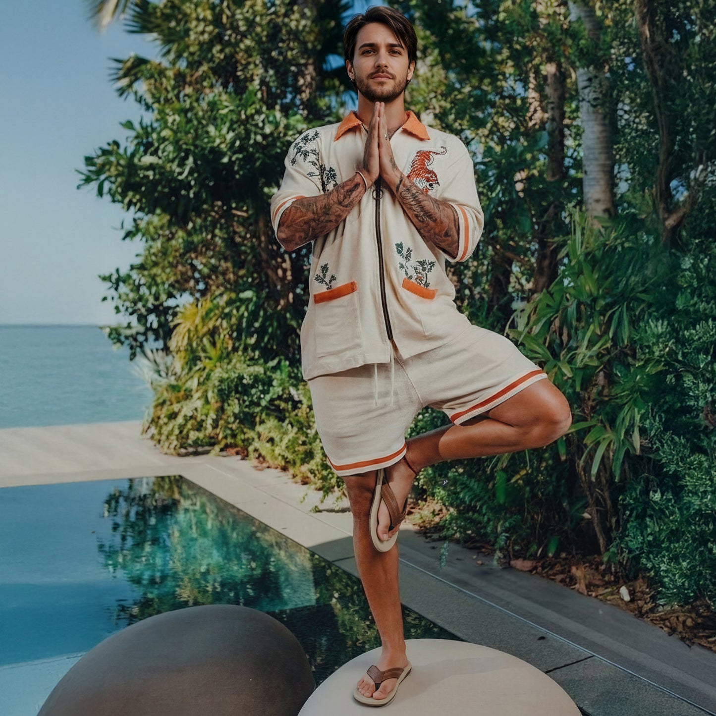 Man in floral embroidered cream shorts and shirt meditating by pool with tropical plants