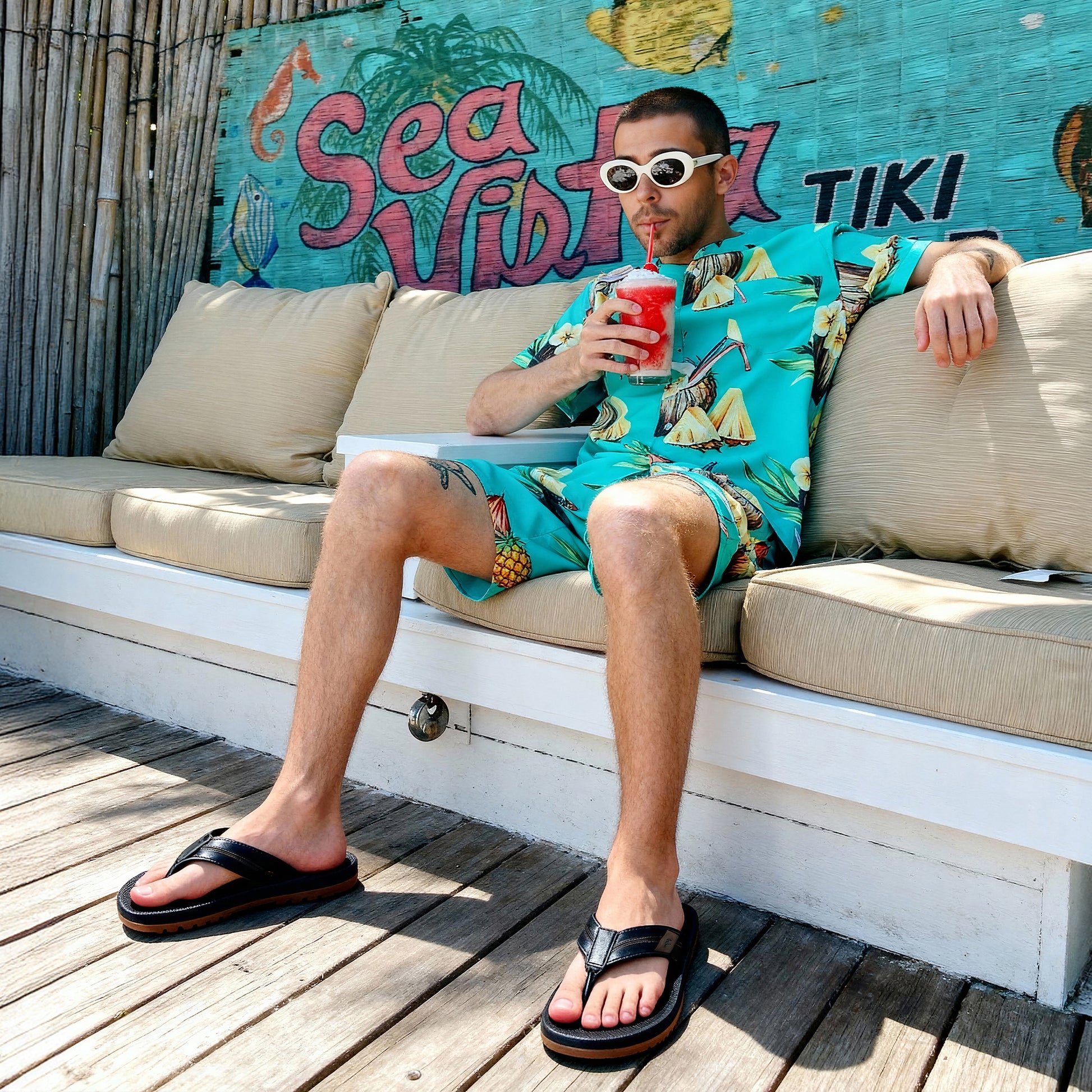 Man in tropical pineapple print outfit and white sunglasses sipping red frozen drink on outdoor patio
