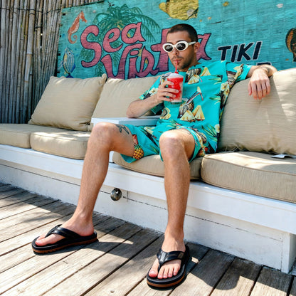 Man in tropical pineapple print outfit and white sunglasses sipping red frozen drink on outdoor patio