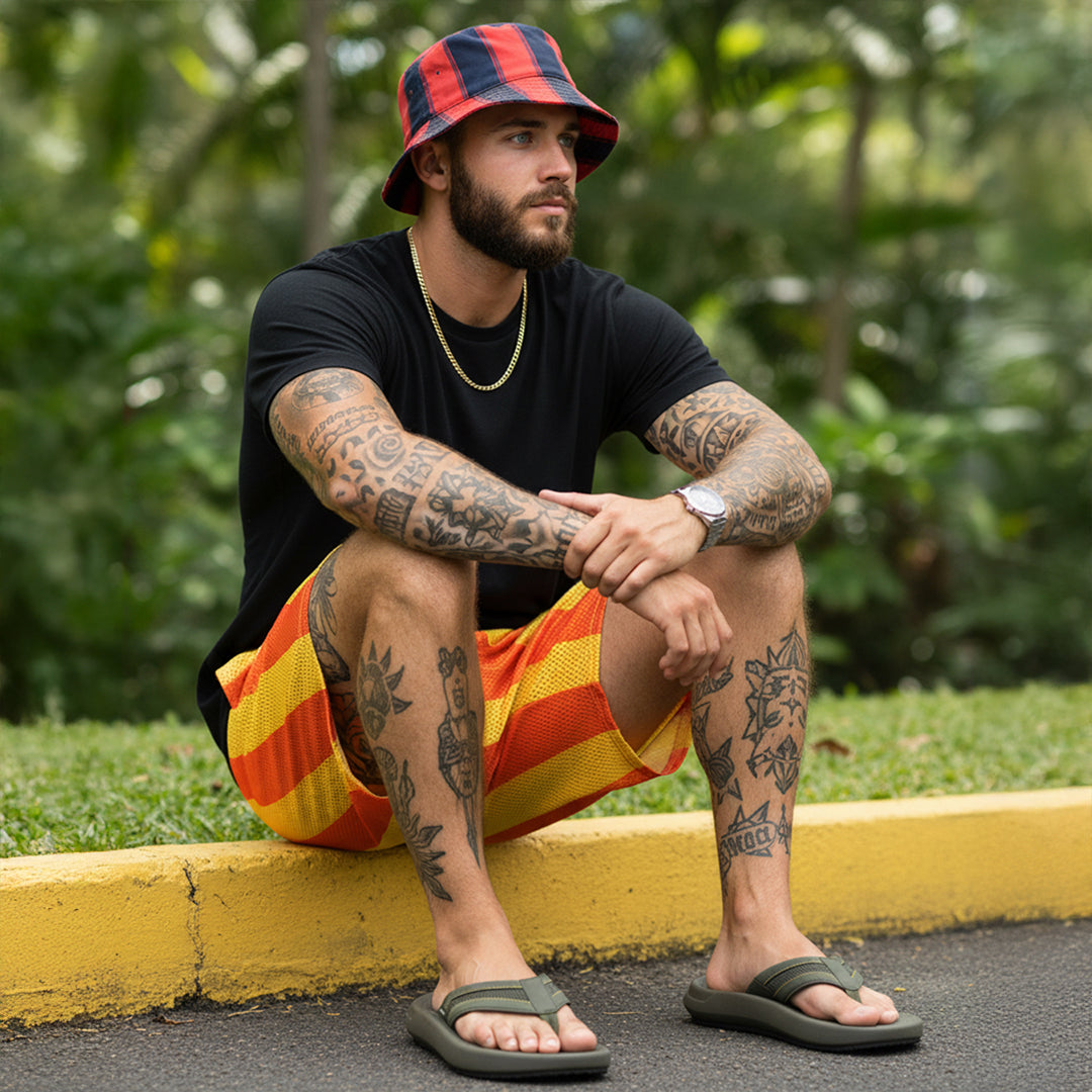 Tattooed man in striped bucket hat, black t-shirt and orange shorts sitting on curb outdoors