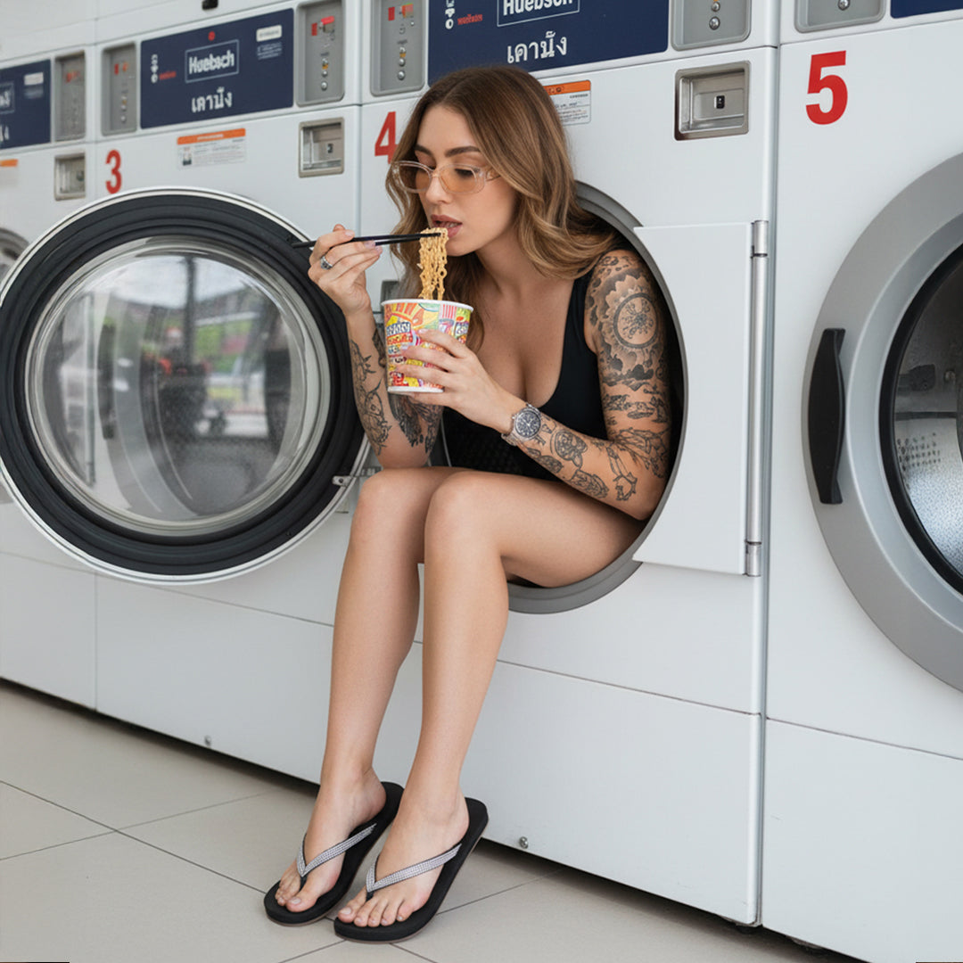 Woman in black flip-flops sitting in laundromat washing machine, eating noodles, tattooed arms