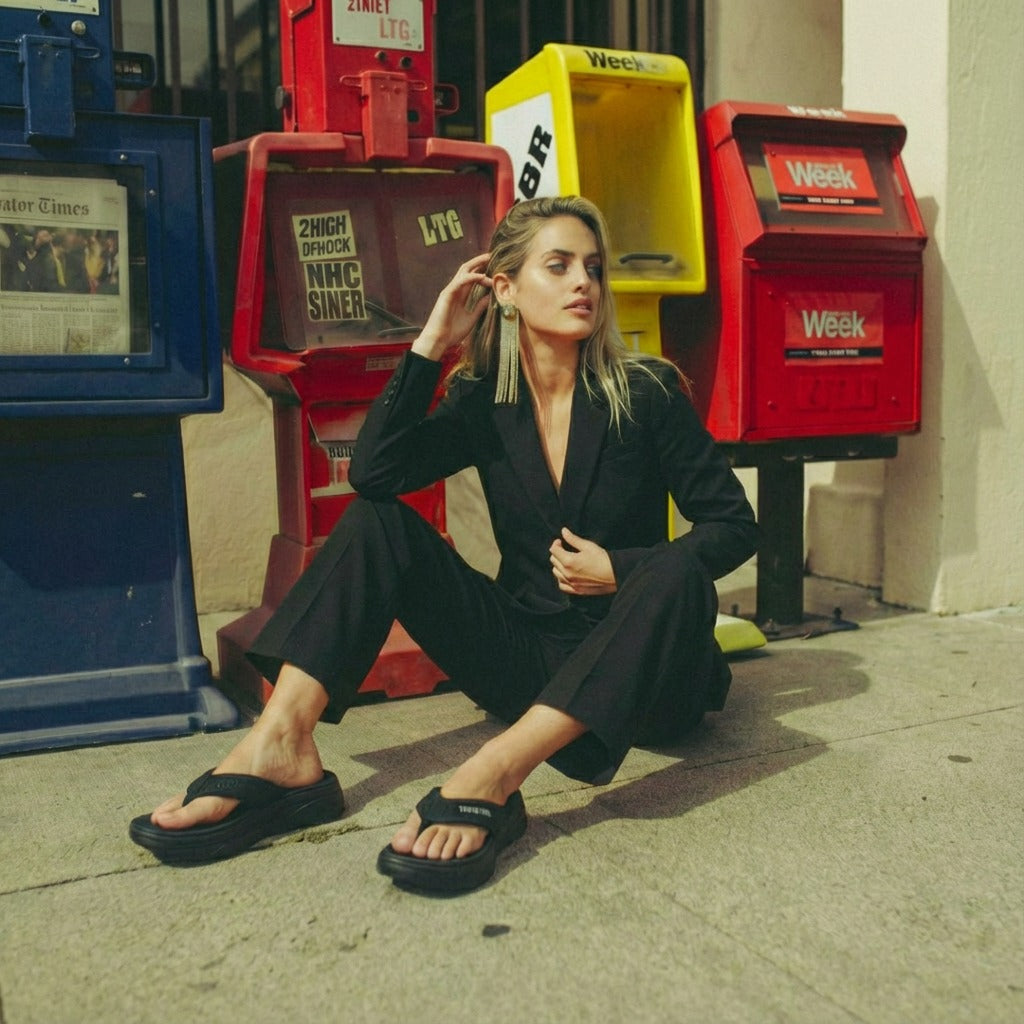 Woman in black suit wearing stylish Toucantoes flip-flops sitting by newspaper stands